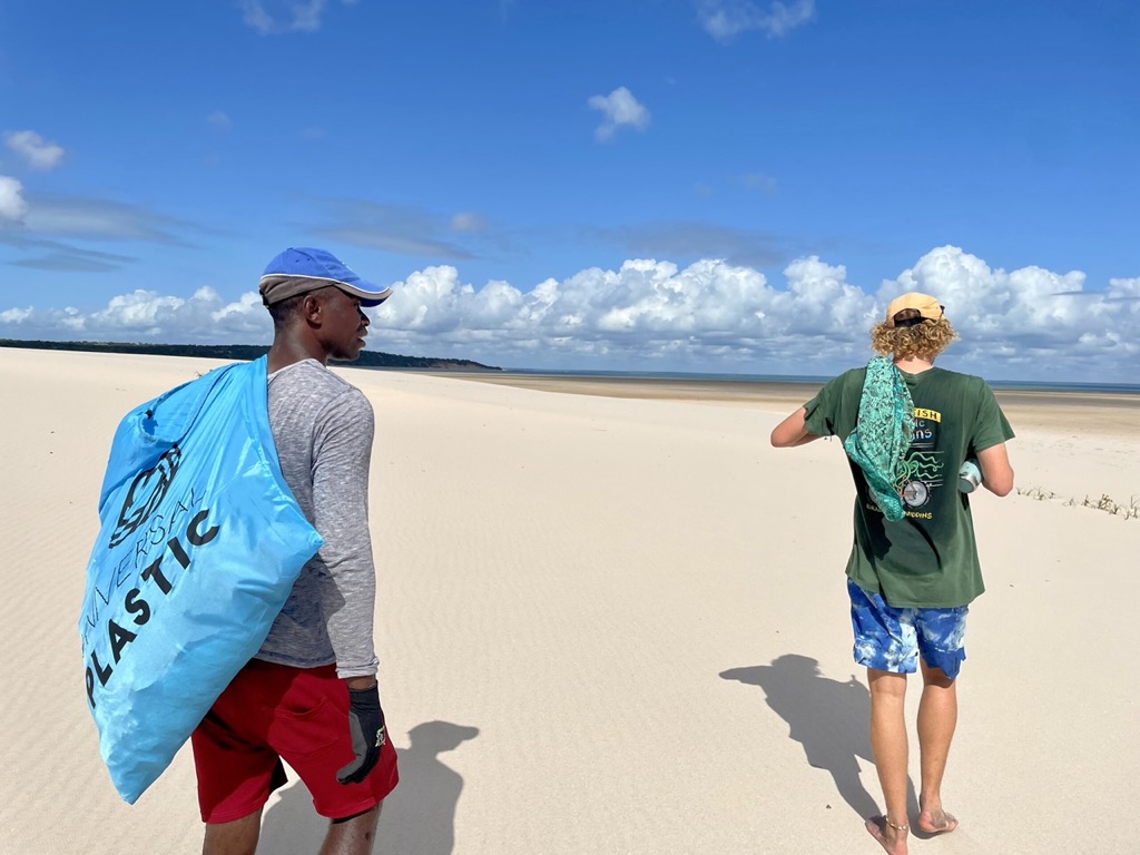 Two people walking along a wide sandy beach, one carrying a large blue Universal Plastic collection bag